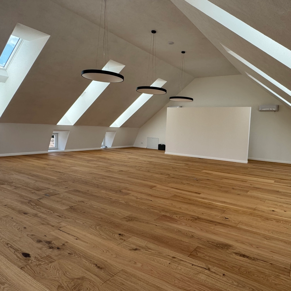 Heller Dachgeschossraum mit Echtholzparkett, modernen Pendelleuchten und Dachfenstern, hochwertig ausgebaut von KraftBau für Wohn- oder Büroflächen.