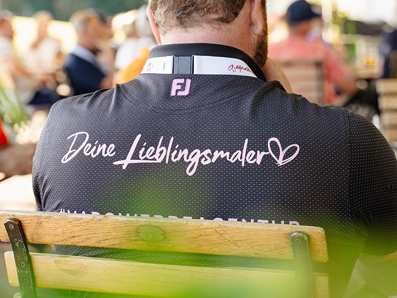 Schwarzes Poloshirt mit rosa Aufdruck „Deine Lieblingsmaler“ und Herz, getragen bei einem Event von KraftBau, stärkt Markenimage und Wiedererkennung.