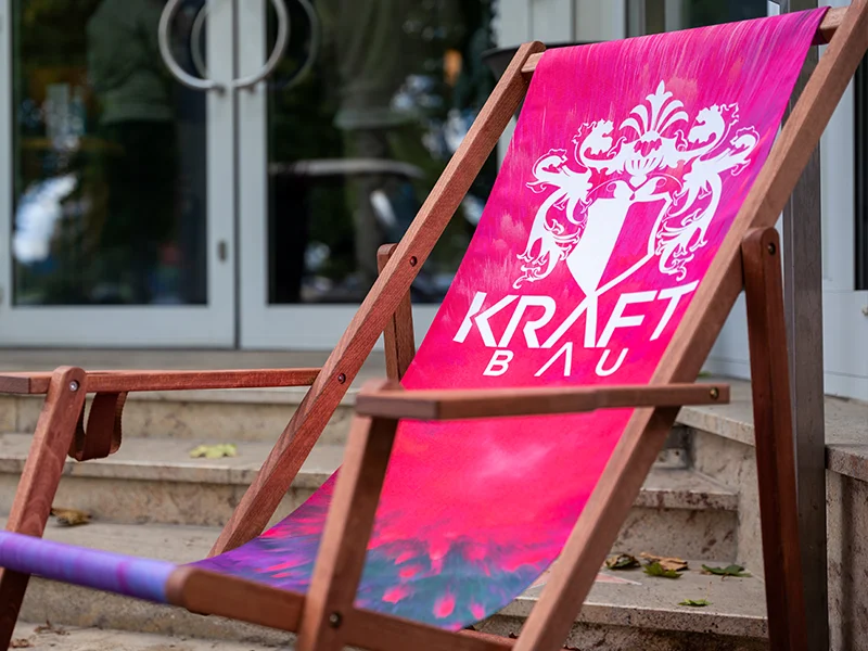 Liegestuhl mit pinkem Stoff und weißem KraftBau-Logo, stilvoll platziert auf Natursteinstufen, perfektes Branding-Element für Firmenevents und Außenbereiche.
