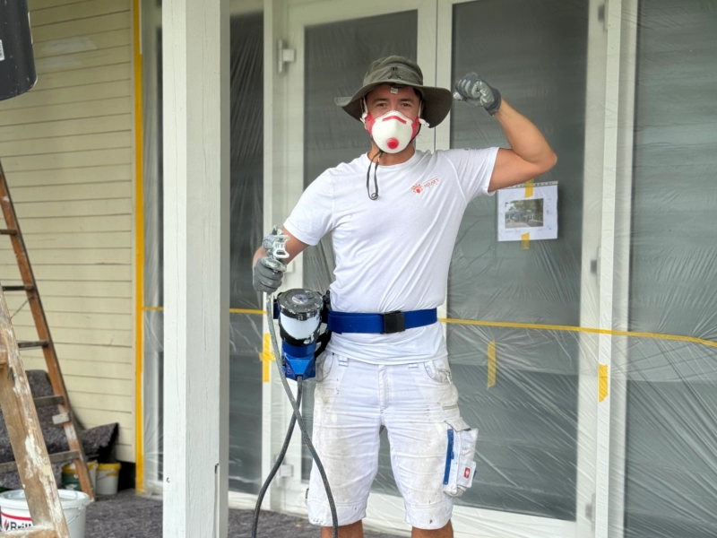 Facharbeiter von KraftBau mit Spritzgerät, Atemschutz und Hut beim Streichen einer Hausfassade, steht für professionelle Malerarbeiten und präzise Ausführung.