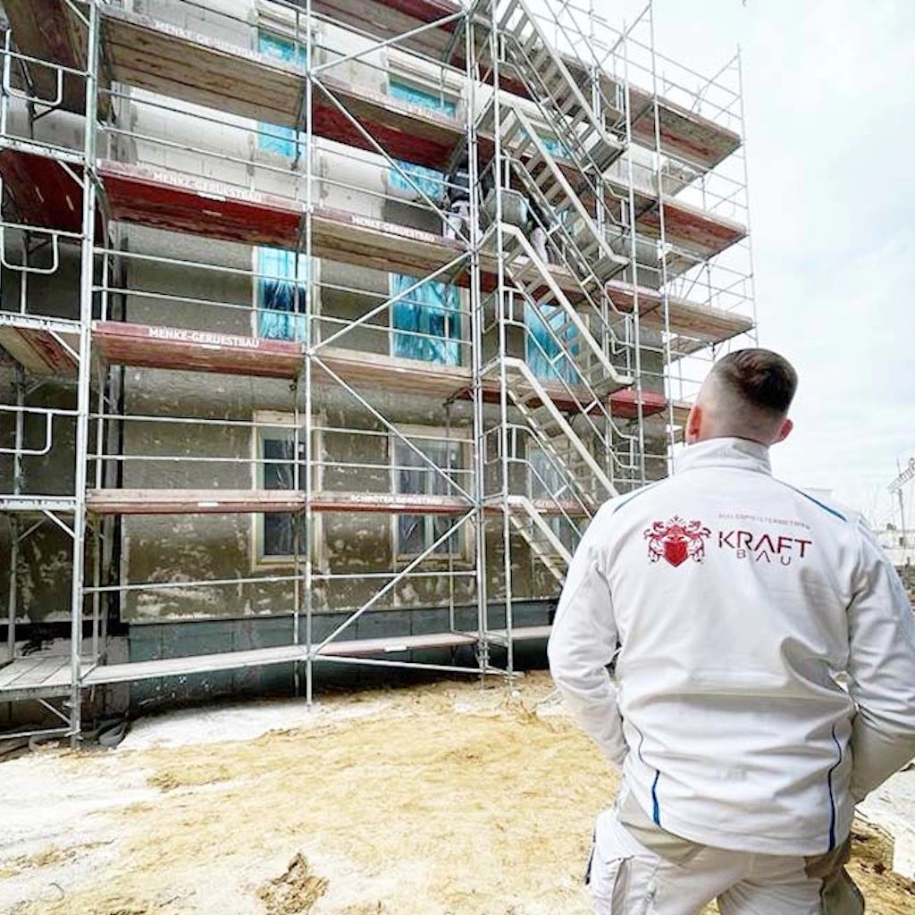 Bauleiter von KraftBau in weißer Arbeitsjacke vor eingerüstetem Mehrfamilienhaus, Symbol für professionelle Fassadensanierung und Projektmanagement.